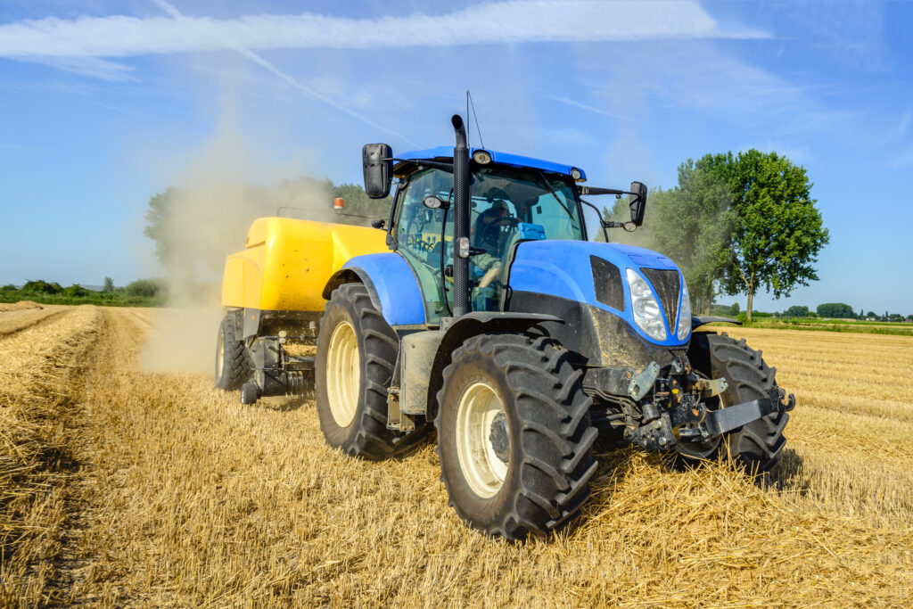 Ein Bauer fährt mit einem Trecker über ein Strohfeld und verarbeitet das Stroh mit einem landwirtschaftlichem Gerät, welches an dem Trecker angebracht ist.