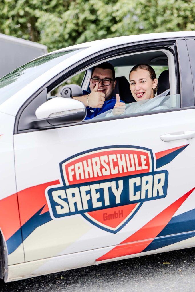 Ein Fahrlehrer und eine Farschülerin in einer Praxisstunde die beide aus dem offenen Fenster der Fahrertür hinaus schauen und und lächelnd einen Daumen hoch zeigen.