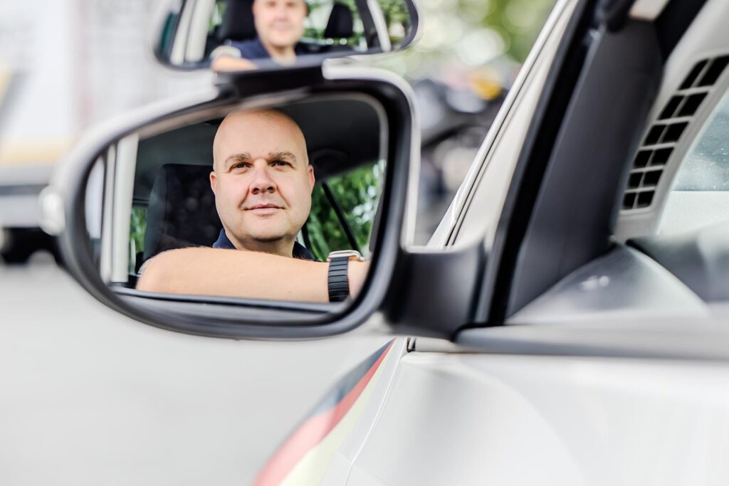 Ein Fahrlehrer der Fahrschule sitzt in einem VW Golf und sein lächelndes Gesicht spiegelt sich im Seitenspiegel des Autos.