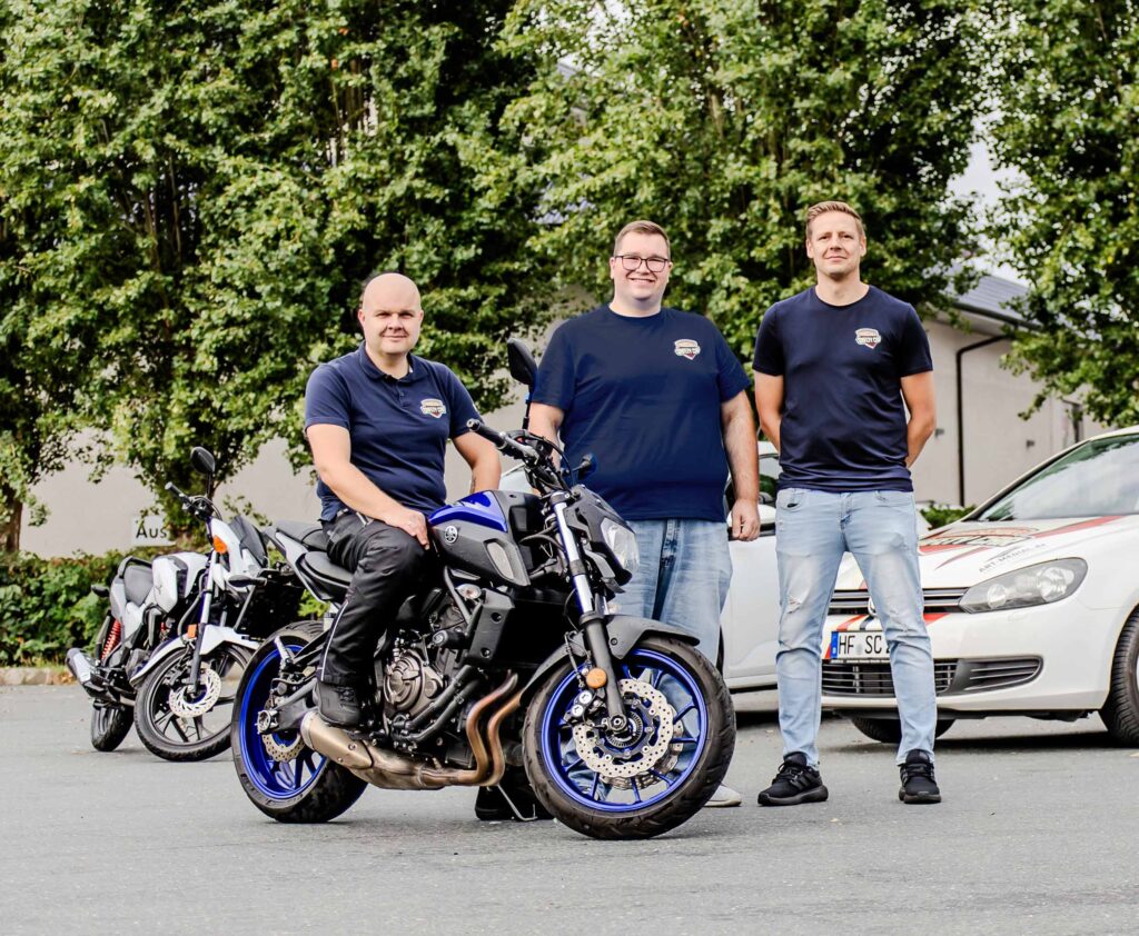 Drei Fahrlehrer im Alter von ca. 34-56 Jahren stehen nebeneinander zusammen auf einem Parkplatz. Der ganz linke sitzt auf einem Motorrad und hinter ihnen Stehen noch einpaar Fahrschulfahrzeuge.