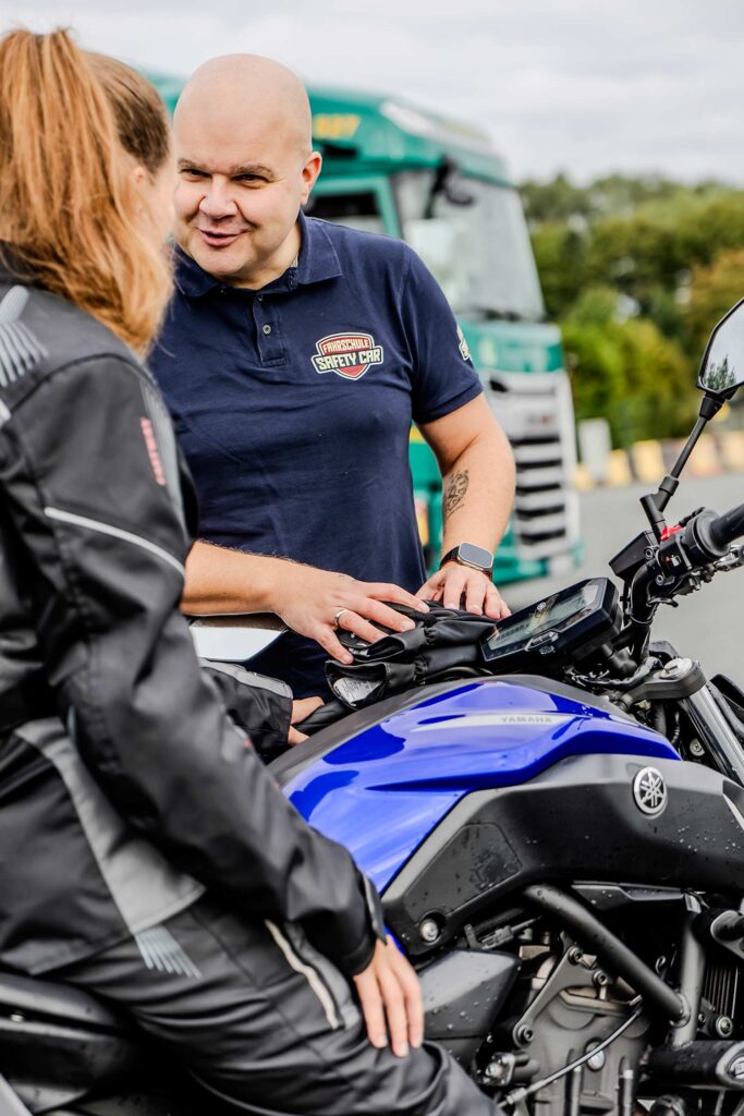 Ein Fahrlehrer erklärt einer Fahrschülerin die auf einem Motorrad sitzt etwas.
