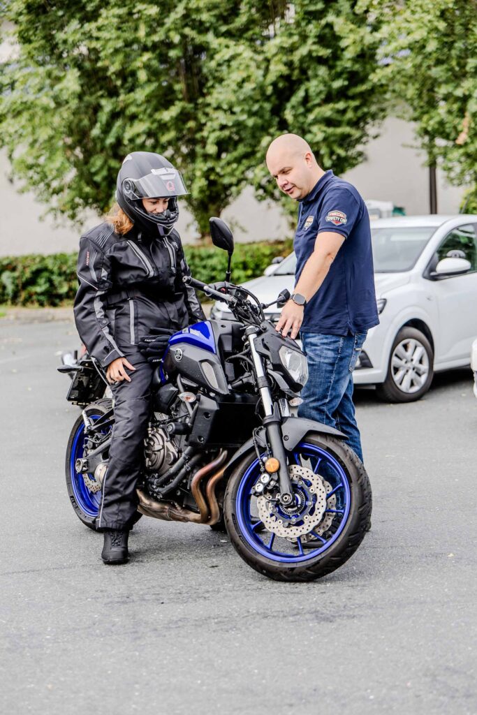 Ein Fahrlehrer erklärt einer Fahrschülerin die auf einem Motorrad sitzt etwas zu den Motorrad Licht Einstellungen.