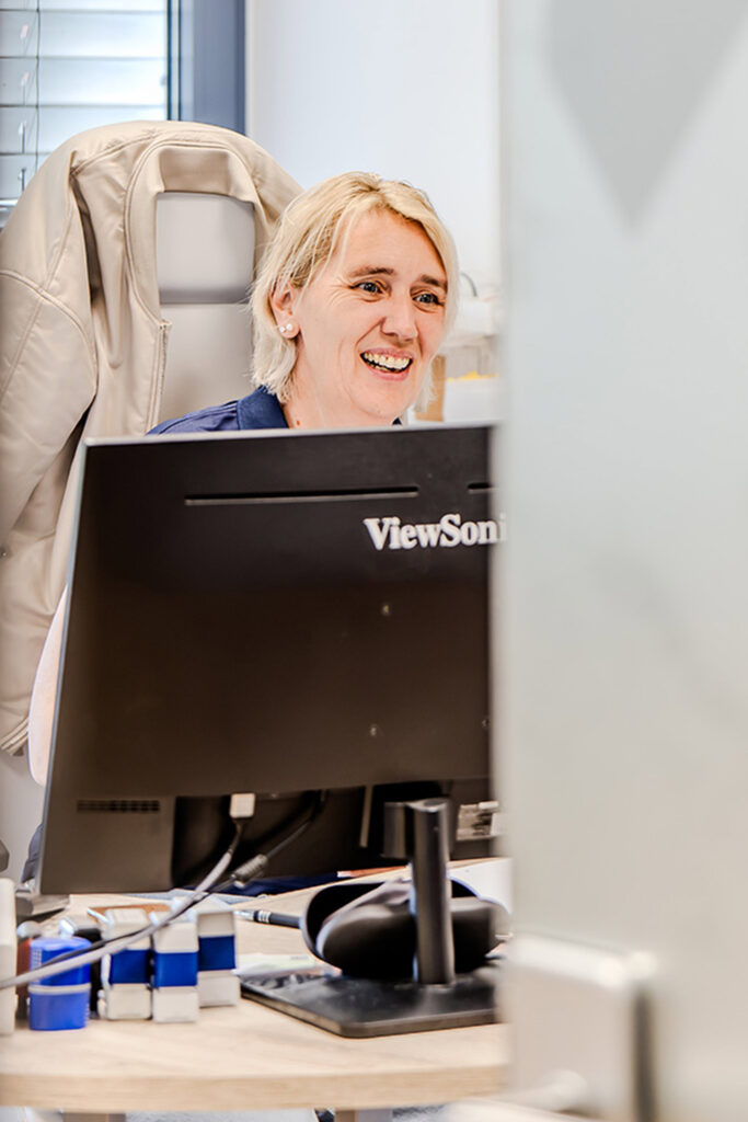Eine Fahrschulmitarbeiterin sitzt im Büro an einem Tisch mit PC und spricht freundlich mit jemandem.
