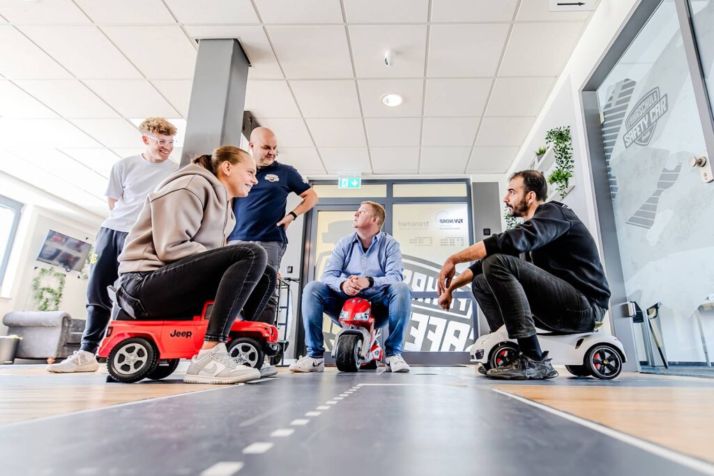Drei Fahrschüler sitzen auf Rutschautos auf einer Straßenkreuzung, die auf dem Flurboden der Fahrschule foliert ist und simulieren eine Verkehrssituation. Der Fahrlehrer erklärt ihnen dazu etwas und die anderen Fahrschüler schauen zu.