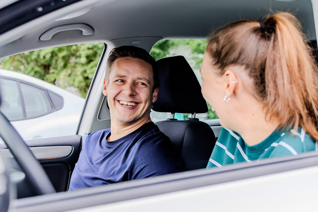 Fahrschülerin und Fahrlehrer sitzen zusammen in einem Fahrschulauto und lächeln sich gegenseitig an. Bild ist von außen durch das offene Fahrerfenster aufgenommen.