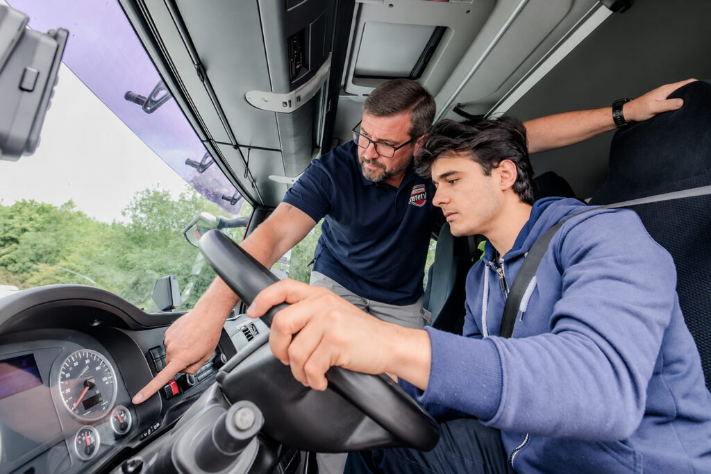 Praxisstunde einer LKW Fahrstunde. Fahrlehrer und Schüler sind im Fahrerraum der LKWs und der Fahrlehrer erklärt dem Fahrschüler alle Anzeigen im Cockpit. Fahrschüler ist im Vordergrund des Bildes und der Fahrlehrer im Hintergrund.