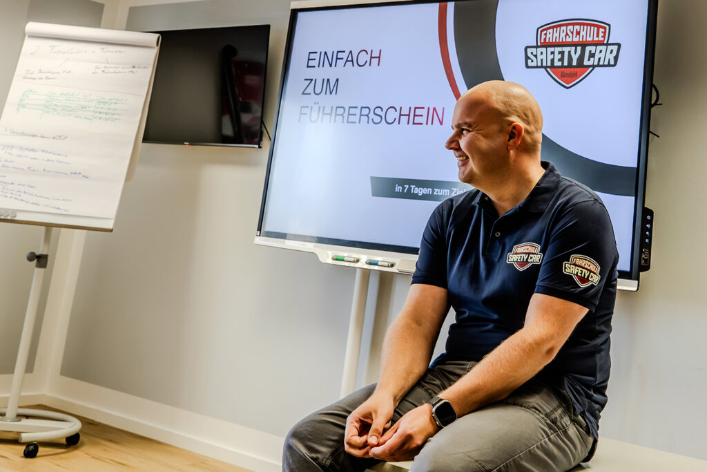 Fahrlehrer sitzt im Therorieunterricht auf einem Tisch. Hinter ihm befindet sich ein Whiteboard und eine Flipchart.